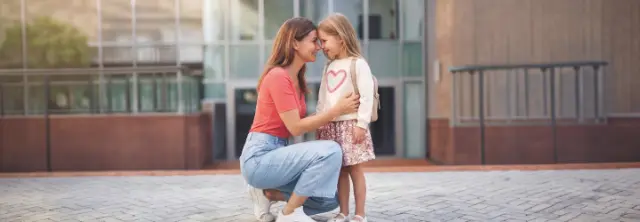 Una madre se despide con cariño de su hija antes de entrar al colegio. La niña lleva una mochila y está lista para comenzar el día de clase.