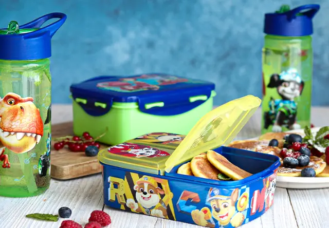 Fiambrera y botellas reutilizables con diseños de PAW Patrol y dinosaurios sobre una mesa preparada con fruta y aperitivos para el colegio o la guardería. Fiambrera y botellas reutilizables con diseños de PAW Patrol y dinosaurios sobre una mesa preparada con fruta y aperitivos para el colegio o la guardería.