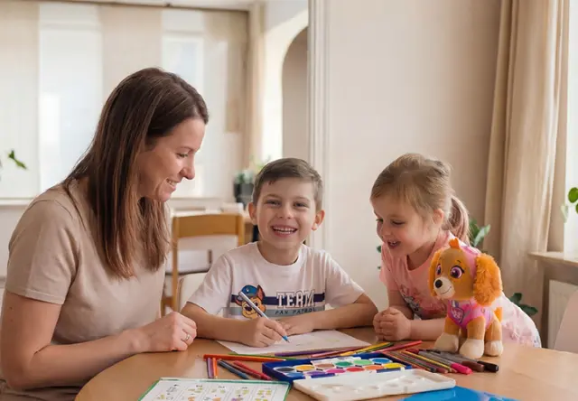 Una mujer pinta junto a dos niños sentados a la mesa. Lápices de colores, acuarelas y un peluche de PAW Patrol invitan a dar rienda suelta a la creatividad. Una mujer pinta junto a dos niños sentados a la mesa. Lápices de colores, acuarelas y un peluche de PAW Patrol invitan a dar rienda suelta a la creatividad.