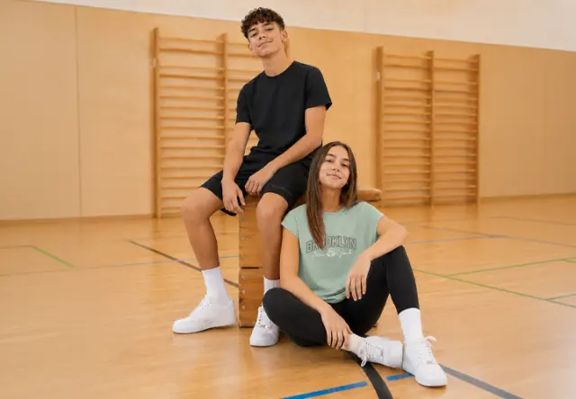 Dos adolescentes llevan ropa deportiva cómoda y están sentados sobre y junto a un potro de gimnasia. Sus outfits son ideales para las clases de educación física.